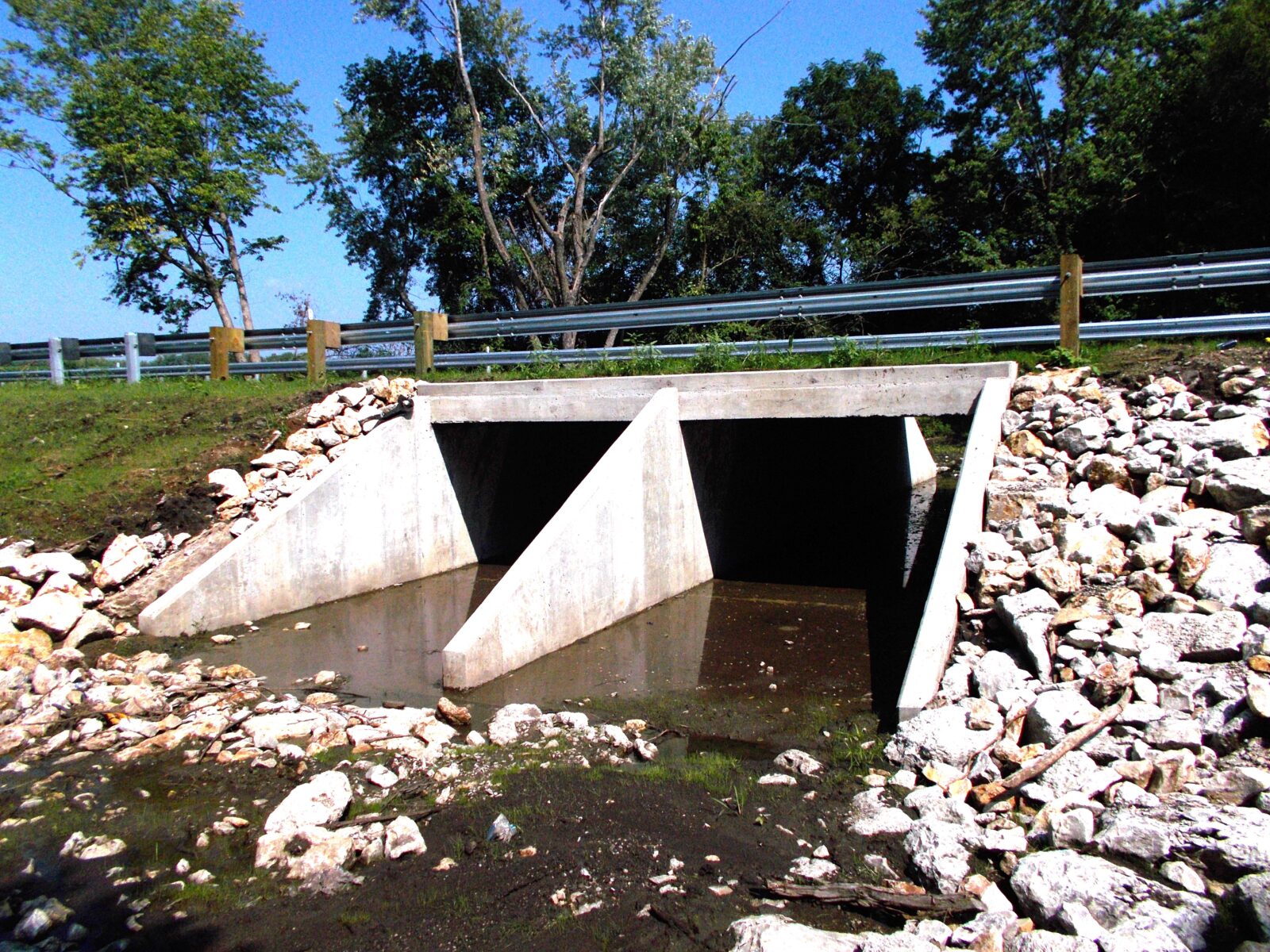An image of a box culvert passing under a bridge. There is riprap on the slopes on either side of the culvert.