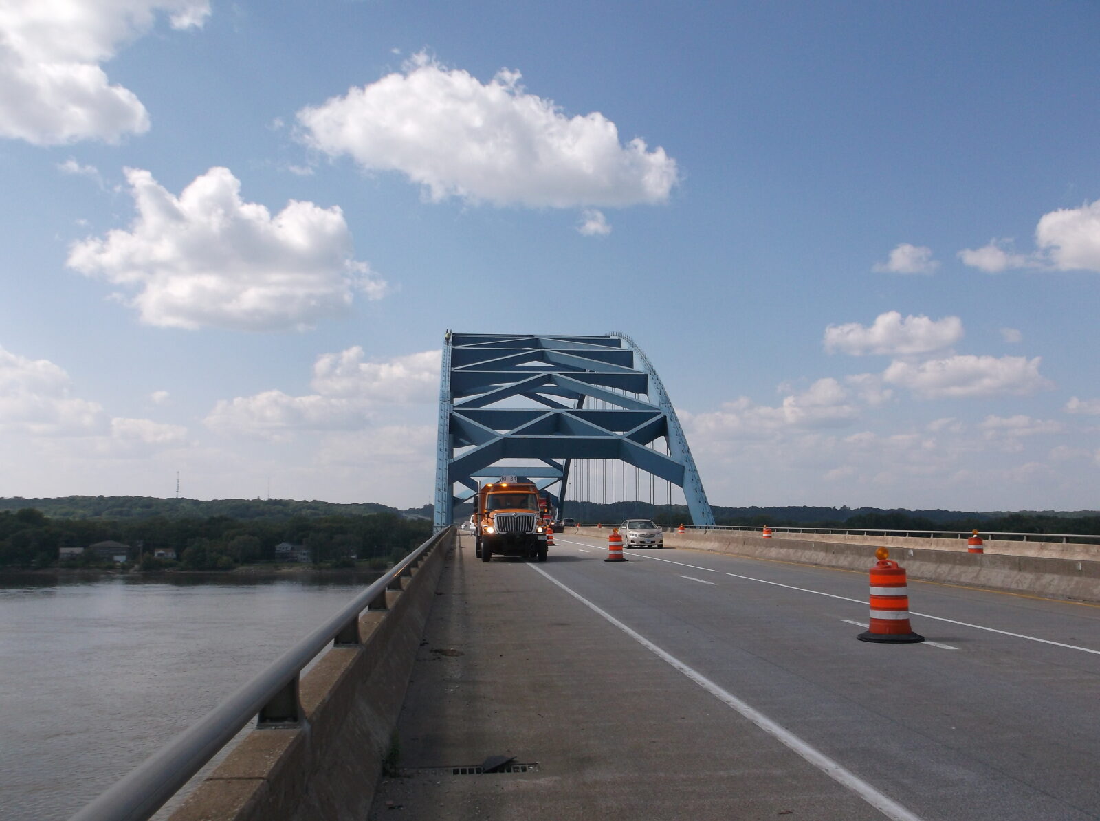 An image of a concrete and steel bridge over a river, taken from the deck of the bridge. Orange barrels are out, indicating there is construction work being performed.