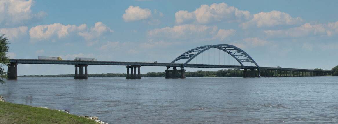 Image of a long steel and concrete bridge over a wide river, taken from the shore.