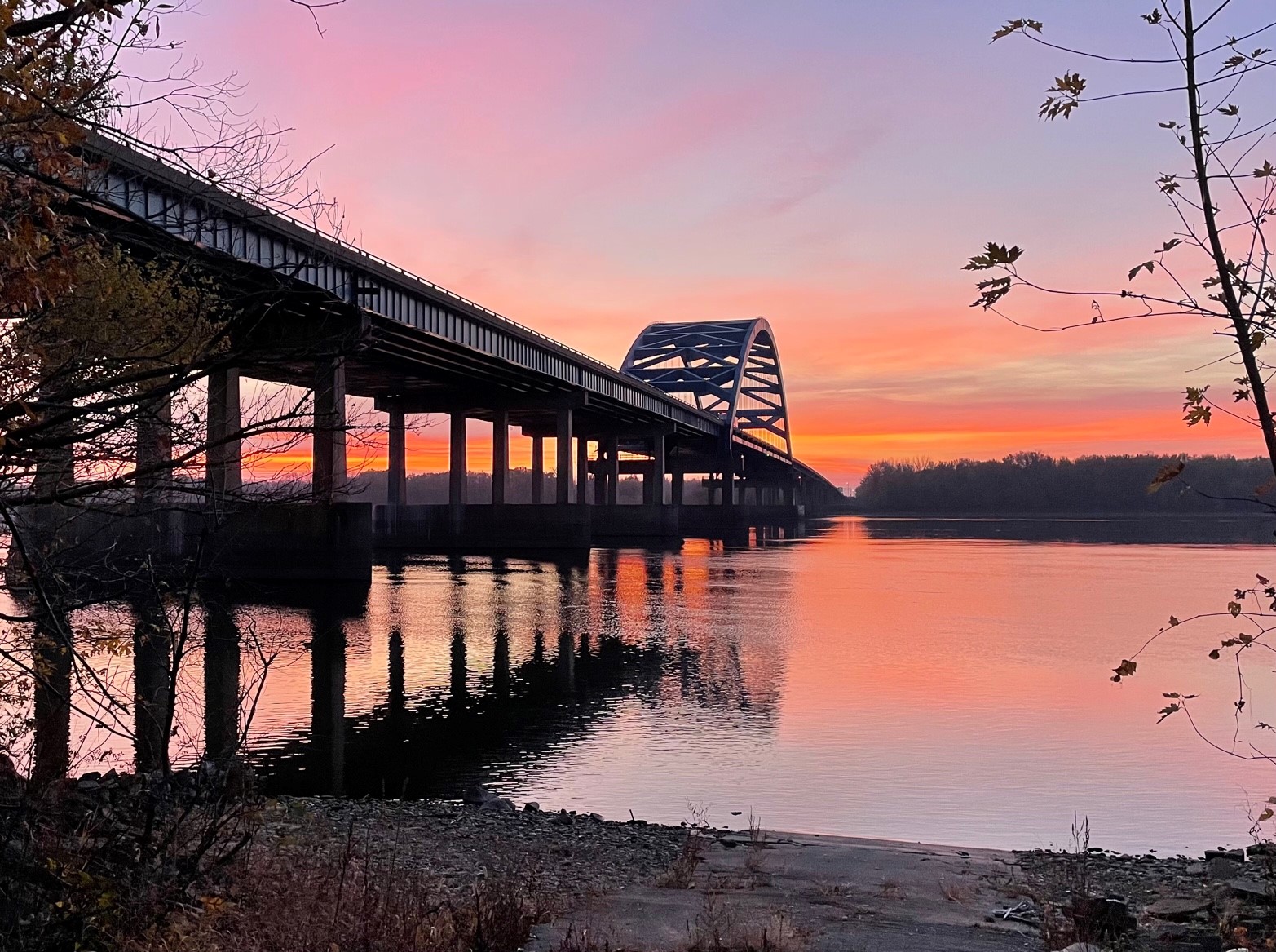 An image of a bridge across a wide river at sunset, taken from the shore.