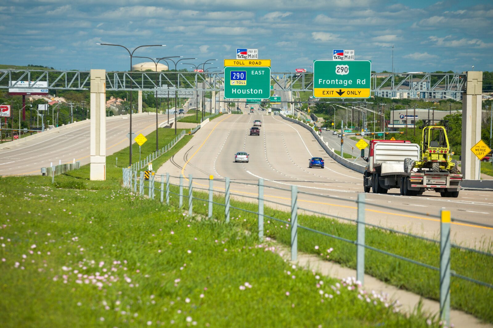 An image of a highway taken from the center median. Two navigational signs on an overhead sign bridge are in focus.