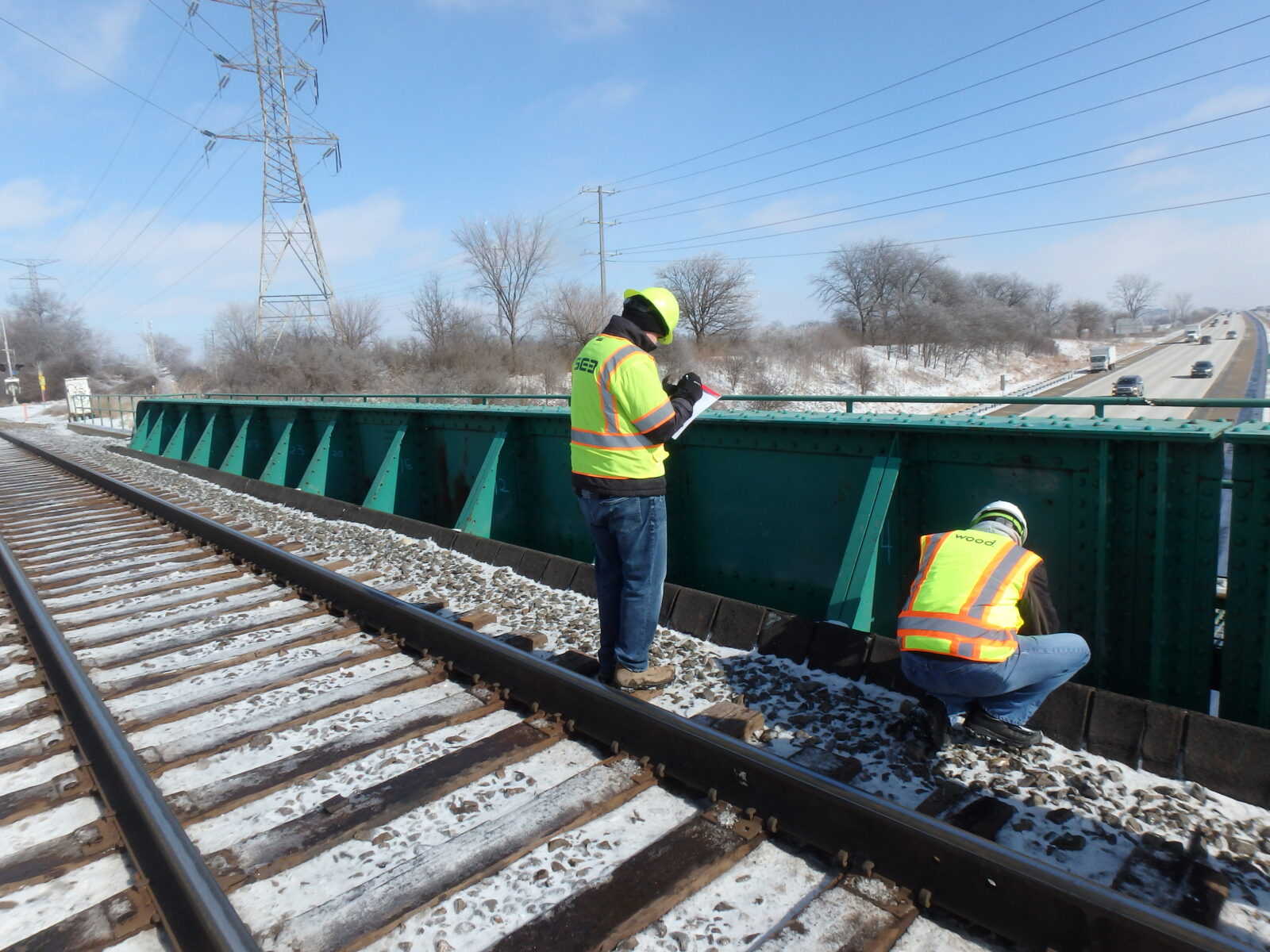 An image of two people in hi-vis clothing inspecting a snowy rail bridge over a highway.