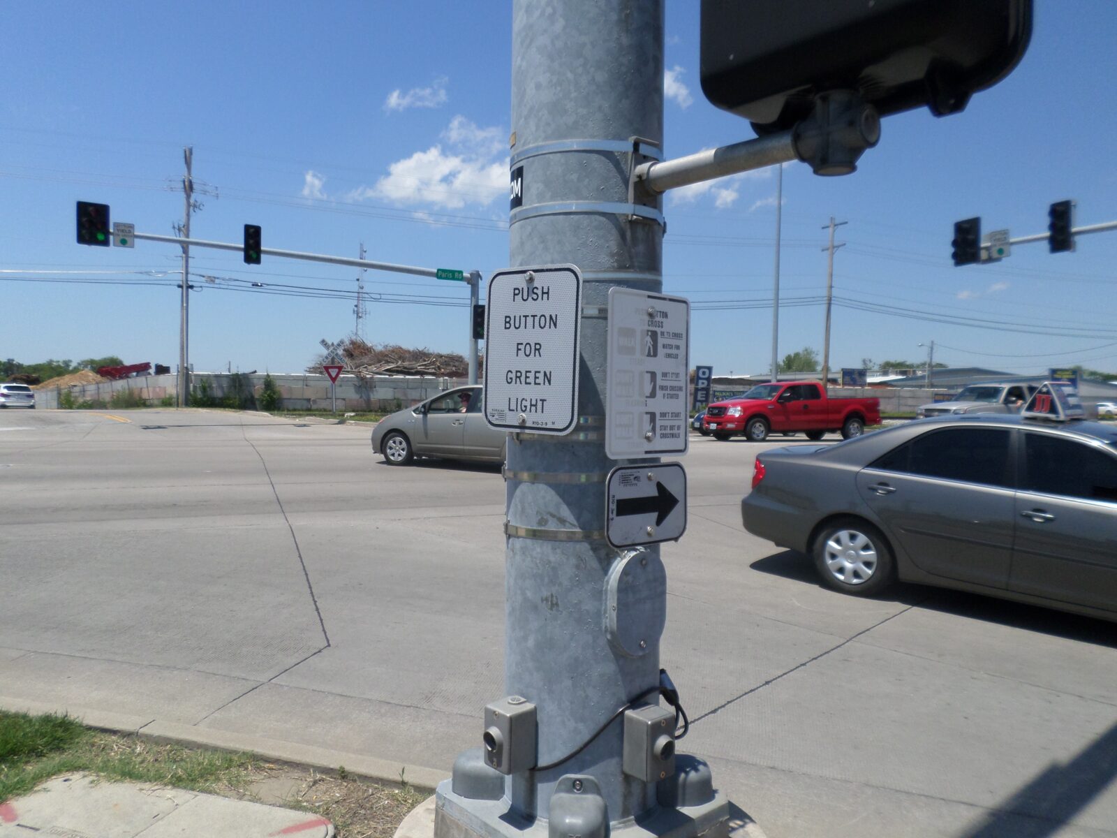An image of a mast arm pole, with a pedestrian signal head attached to it, as well as signs instructing pedestrians on requesting a green phase and crossing the street.