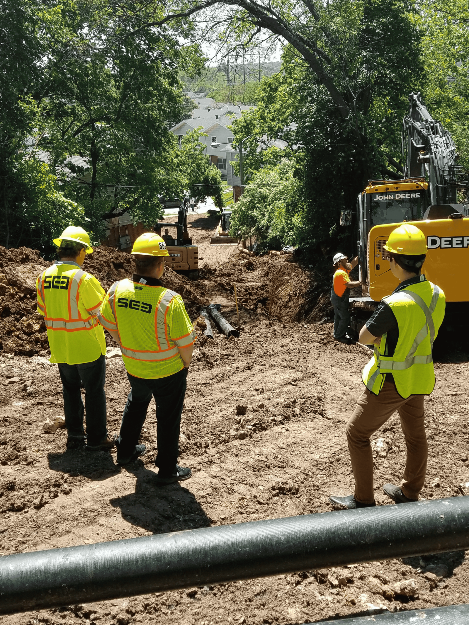 Image of a construction site, with excavating equipment and people in hi-vis vests with "SE3" on the back. They're looking at an excavated ditch with large pipes in it.