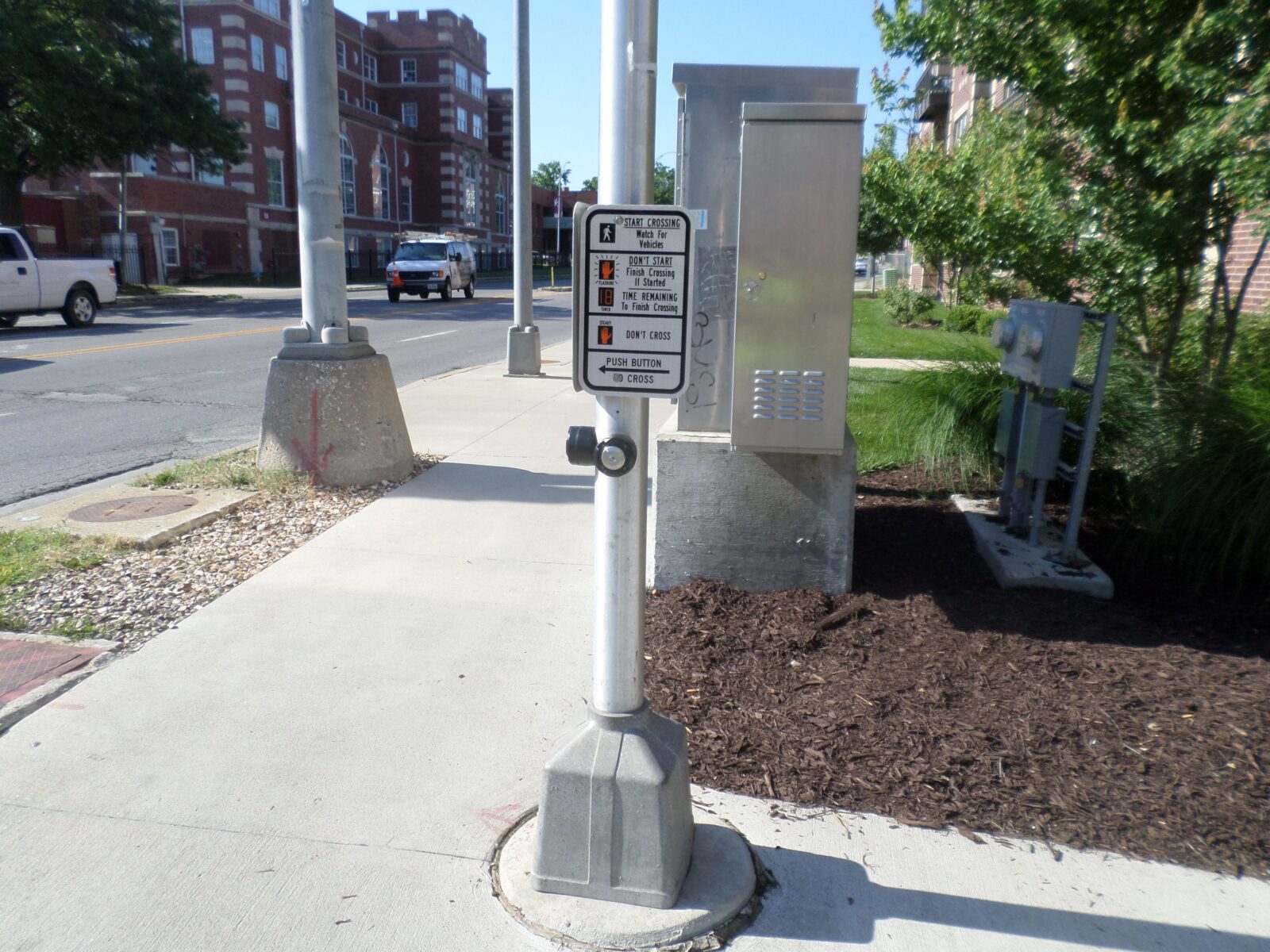 An image of a pole with two pedestrian push-buttons on it. Both push-buttons are accompanied by a sign instructing pedestrians on crossing the street.