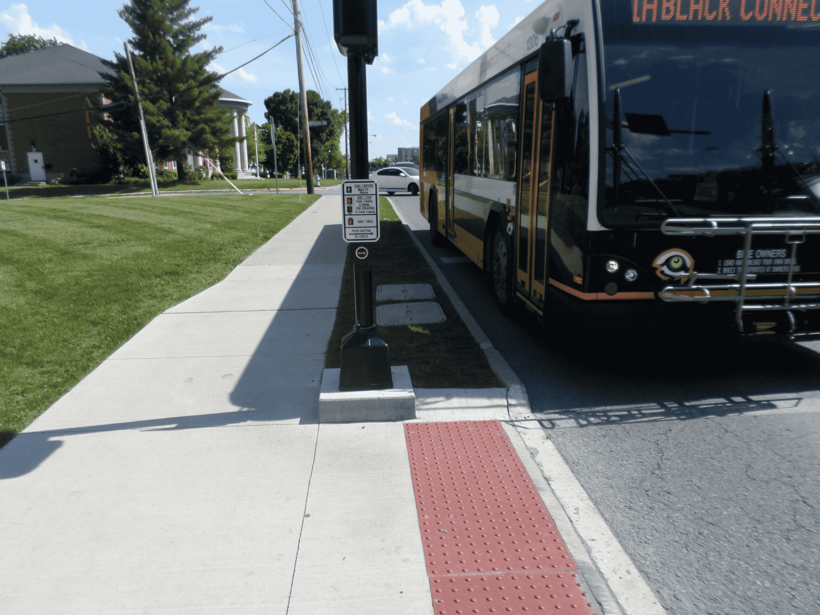 An image of a pedestrian push button on a signal post at an intersection. There is a bus next to the signal pole.